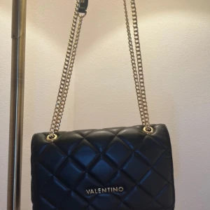 Svart quiltad axelväska från Valentino - Snygg svart axelväska från Valentino med quiltad design och guldfärgade kedjor. Väskan är tillverkad i skinn och har ett elegant metallspänne framtill samt logga i guld. Perfekt storlek för det viktigaste och ger en lyxig känsla till din outfit.