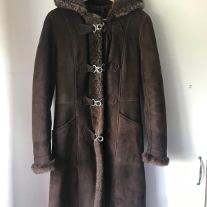 Brun lång pälsjacka med huva - Snygg lång brun pälsjacka med huva och pälsdetaljer vid ärmslut och insida. Jackan har dekorativa spännen framtill, stora fickor och bälte i ryggen. Perfekt för kalla dagar när du vill hålla stilen och värmen.