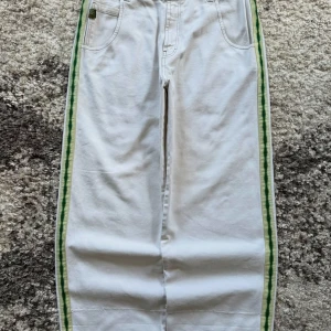 JNCO vita wide jeans med gröna ränder - Unika vita jeans från JNCO med bred passform och gröna ränder längs sidorna. Bakfickan har en broderad grön orm med krona. Jeansen är tillverkade i 100% bomull och har coola detaljer som gröna sömmar och klassiska fem fickor.                                                             mått 91 cm midja 89 cm innerbenssöm 112 cmv yttersöm 63 cm benöppning/28 cm platt