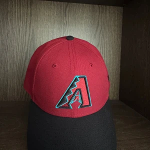 Arizona Diamondbacks keps New Era - Röd och svart keps från New Era med Arizona Diamondbacks logga broderad framtill och justerbar rem baktill. Kepsen har böjd skärm och klassisk basebollstil, perfekt för dig som gillar sportig look och vill representera ditt lag.