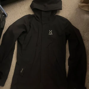 Svart regnjacka från Haglöfs med huva - Svart regnjacka från Haglöfs med stilren design och huva. Jackan har diskret logga på bröstet och dragkedja framtill. Tillverkad i slitstarkt syntetmaterial som står emot regn och vind. Perfekt för dig som vill ha en clean look även när det är dåligt väder.