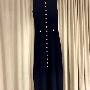 Marinblå långklänning med guldknappar - Snygg marinblå långklänning med ärmlös design och raka linjer. Klänningen har dekorativa guldknappar längs hela framsidan och två fickdetaljer vid midjan. Materialet är stickat och stretchigt, vilket ger en bekväm passform.