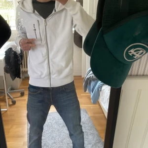 Vit hoodie från Sail Racing med dragkedja - Vit hoodie från Sail Racing med hel dragkedja framtill och tvär fet att ha på sig men också att ha under en jacka, det blir skit snyggt! Broderad logga på vänster sida. Tröjan har huva och ribbade muddar vid ärmslut och nederkant. Perfekt för en clean och sportig stil.finns lite fläckar men går nog bort i tvätten.