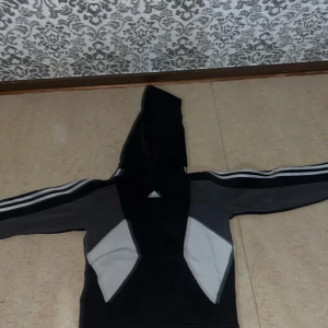 Svart och grå Adidas hoodie - Säljer en snygg hoodie från Adidas i svart och grått med vita detaljer. Tröjan har huva, långa ärmar med klassiska Adidas-ränder och en stor ficka framtill. Perfekt för dig som gillar sportig stil och vill ha något bekvämt och coolt.