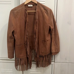 DAY Birger et Mikkelsen mockajacka med fransar  - Säljer en rosa mockajacka från DAY Birger et Mikkelsen i getskinn. Jackan har långa ärmar, öppen front och coola fransdetaljer längs kanterna och nederkanten. Perfekt för dig som vill sticka ut med en boho-vibe.