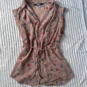 Beige blommig blus från Bik Bok - Supersöt beige blus från Bik Bok med små blommor i rött, blått och grönt. Blusen är ärmlös, har knappar framtill, v-ringning och ett knytband i midjan för en snygg passform. Perfekt för varma dagar och enkel att styla med jeans eller kjol.