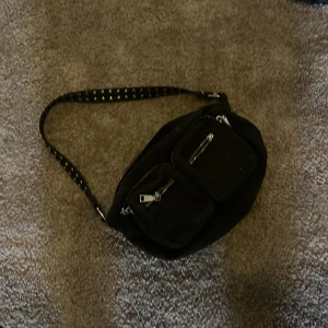 Svart axelväska med nitar och dragkedjor - Cool svart axelväska med bredt axelband dekorerat med silvriga nitar. Väskan har flera praktiska fack framtill med dragkedjor, perfekt för att hålla ordning på dina grejer. Stilren och edgy design som passar till många outfits.