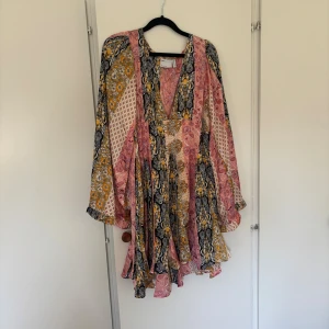 Bohemisk kortklänning  - Säljer en bohemisk kortklänning med v-ringning och volangkant. Klänningen har ett färgstarkt paisleymönster i rosa, gul och svart, samt långa, vida ärmar. Perfekt för dig som gillar unika och färgglada plagg! Ganska kort i längd