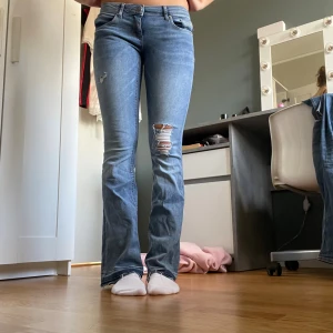 Blå bootcut jeans från ONLY - Snygga blå bootcut jeans från ONLY, modell CORAL. Jeansen har låg midja, slitningar och hål på ena benet samt råa, fransiga benslut. Klassisk femficksmodell i stretchig denim som sitter tajt upptill och släpper ut vid vaden.