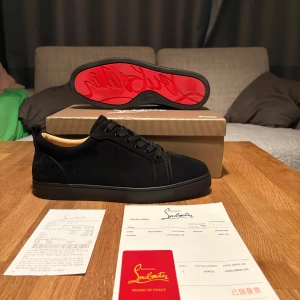 Christian Louboutin svarta sneakers - Svarta sneakers från Christian Louboutin i mocka med klassisk röd sula och diskret broderad logga på hälen. Snygg och stilren design med snörning och rund tå. Insidan är ljusbeige och skon har en platt sula. Perfekt för dig som vill sticka ut med exklusiva detaljer.
