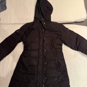 Svart lång pufferjacka från Sebago - Svart lång pufferjacka från Sebago med huva, dragkedja och tryckknappar framtill. Jackan har quiltad design, fickor med dragkedja och en liten ficka på ärmen. Perfekt för kalla dagar och har en klassisk look. Finns tyvärr ett hål som finns bild på men de är väldigt litet. Pris går att diskutera 