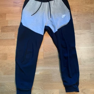 Nike Tech byxor - Nike mjukisbyxor i svart och ljusgrått med vit logga på vänster sida. Byxorna har snörning i midjan, muddar vid bensluten och en dragkedjeficka bak. Tillverkade i mjukt bomullsmaterial. Liten slitning på baksidan. Skriv vid minsta lilla fundering!