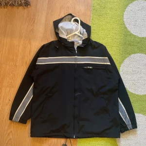 Svart vindjacka från Superpros - Svart vindjacka med huva från Superpros Sportswear. Jackan har gråa och vita reflexdetaljer längs bröstet och ärmarna, meshfoder på insidan och dragkedja framtill. Praktiska fickor och justerbar nederkant med snörning. Perfekt för blåsiga dagar.