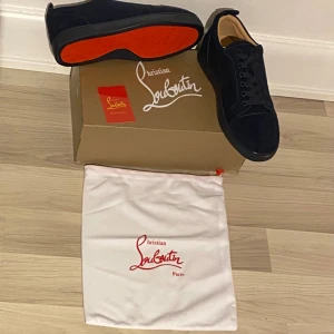 Christian Louboutin marinblå mockasneakers - Exklusiva sneakers från Christian Louboutin i marinblå mocka med ikonisk röd sula. Skorna har rund tå, snörning och diskret broderad logga på hälen. Insidan är fodrad i beige skinn för extra komfort. Perfekt för dig som vill sticka ut med lyxiga detaljer.