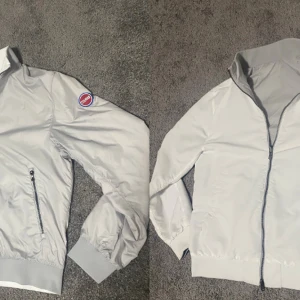 Unik vindjacka från Colmar - Dubbelfärgad vindjacka från Colmar med hög krage, svarta dragkedjor och ribbade muddar. Jackan har en klassisk passform, lång ärm och en diskret logotyp på ärmen. Perfekt för blåsiga dagar och enkel att matcha med det mesta. Går att ha som både vit och grå. Hör av er vid fler frågor😁