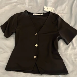 Svart blus med v-ringning och knappar - Svart blus med v-ringad hals och tre snygga knappar framtill. Modellen har korta ärmar och en lätt figursydd passform. Perfekt att styla med jeans eller kjol för en clean look.