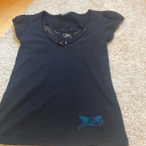 Svart t-shirt Rock De Lux med spetsdetaljer - Svart t-shirt från Rock De Lux med coolt blått tryck nertill och snygga spetsdetaljer vid v-ringningen och på ryggen. Kortärmad modell i mjukt bomullstyg, perfekt för dig som gillar rockig stil och unika detaljer.