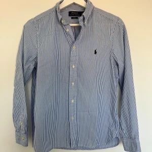 Blårandig skjorta från Polo Ralph Lauren - Snygg blårandig skjorta från Polo Ralph Lauren i slim fit-modell. Skjortan har klassisk krage, långa ärmar med manschett och knappar framtill. Tillverkad i mjuk bomull och har den ikoniska broderade loggan på bröstet. Perfekt för en clean och fräsch look.