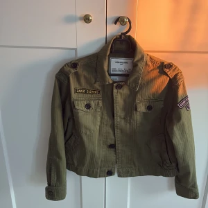 Olivgrön jacka från Zadig & Voltaire - Säljer en olivgrön jacka från Zadig & Voltaire med coola badges och knappar på axlarna. Jackan har två bröstfickor med lock och knappar, samt detaljerade sömmar. Perfekt för dig som gillar en avslappnad men edgy stil.