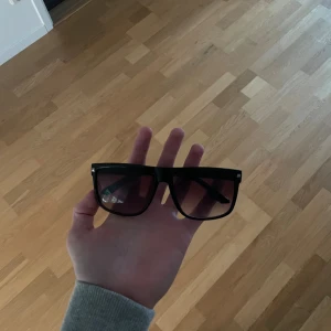 Svarta fyrkantiga solglasögon - Säljer ett par stilrena svarta solglasögon med breda bågar och fyrkantig form. Glasen är mörka med en lätt toning som går från svart till brunt. Perfekta för att ge en cool vibe till din outfit.