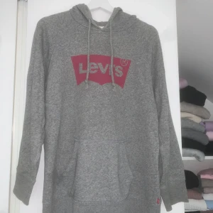Grå hoodie från Levi's med logga - Grå hoodie från Levi's med klassisk röd logga framtill. Tröjan har huva med snörning, magficka och långa ärmar.