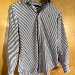 Randig skjorta från Polo Ralph Lauren - Snygg skjorta från Polo Ralph Lauren i slim fit med blå och vita vertikala ränder. Klassisk krage, långa ärmar och knappar framtill. Stretchigt bomullsmaterial för skön passform. Ikonisk broderad logga på bröstet. Perfekt för dig som gillar stilrena och fräscha plagg.