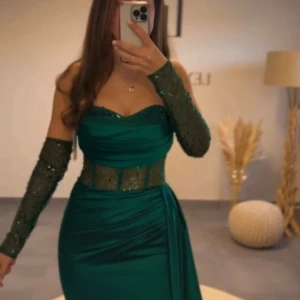 Grön långklänning med glitterdetaljer - Snygg grön långklänning med offshoulder och glittriga meshdetaljer vid midjan och på ärmarna. Klänningen har en figurnära passform och ett elegant fall. Köpt i tyskland, använt 1 gång