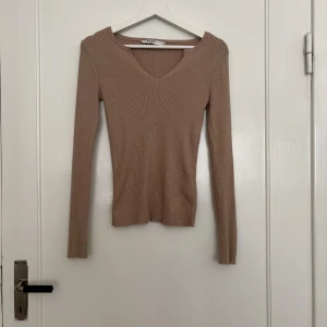 Beige ribbad långärmad tröja NA-KD - Säljer en beige ribbad långärmad tröja från NA-KD med v-ringning. Tröjan har en slim passform och är tillverkad i mjukt, stretchigt material som sitter snyggt på kroppen. Perfekt att styla med jeans eller kjol.