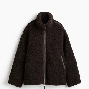 Svart teddyjacka med dragkedja - Mysig svart teddyjacka med hög krage och grov dragkedja framtill. Jackan har två stora fickor och detaljer i syntet längs ärmarna. Perfekt oversized passform för en chill look och extra värme under kalla dagar.