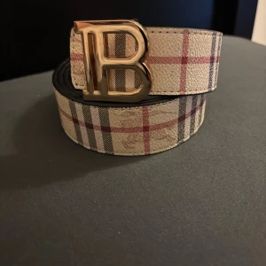 Burberry vändbart bälte med B-spänne - Snyggt vändbart bälte från Burberry med klassiskt rutigt mönster i beige, svart och rött på ena sidan och svart präglat rutmönster med Burberry-ryttare på andra sidan. Stort guldfärgat B-spänne framtill. Perfekt accessoar för att lyfta din outfit. Kom med bud