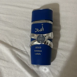 Lattafa Asad Zanzibar parfym blå - Snygg parfymflaska från Lattafa i en djupblå färg med silverdetaljer och orientaliska inslag. Flaskan har en modern design med arabiska tecken och texten Asad Zanzibar. Perfekt för dig som gillar unika dofter och coola flaskor.