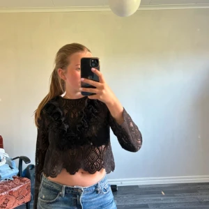 Svart spetsblus med volangdetaljer - Säljer en svart, croppad spetsblus med långa ärmar och fina volangdetaljer framtill. Blusen har ett genombrutet spetsmönster och vågig kant nertill. Perfekt att styla med jeans eller kjol för en trendig look.