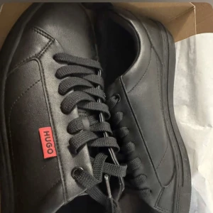  Hugo Boss Herr Morris Faux leather skor  - Sneakers från Hugo Boss med en stilren design och diskret röd logotyp på sidan samt på innersulan. Skorna är en returnerad outletvara och är i mycket gott skick. Ovandelen och insidan ser nästintill nya ut, medan yttersulan visar mindre spår som tyder på att de kan ha provats eller använts vid ett enstaka tillfälle. Paket medföljer och snabb leverans! 