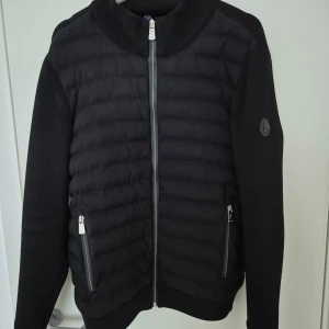 Svart jacka från Moncler - Svart jacka från Moncler med quiltad framsida och stickade ärmar. Jackan har två fickor med dragkedja, ribbad krage och Moncler-logga på ärmen. Snygg och stilren design. 