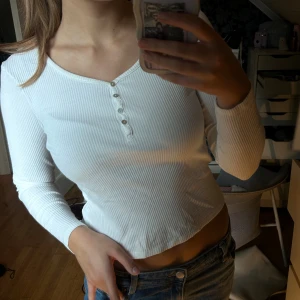 Vit ribbad långärmad topp med knappar - Säljer en vit ribbad långärmad topp med v-ringning och fyra små knappar framtill. Toppen är croppad och har en slim passform som sitter snyggt på kroppen. Perfekt att matcha med jeans eller kjol för en clean och fräsch look.