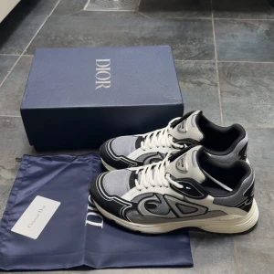 Dior B30 sneakers svart/vit/grå - Säljer ett par Dior B22 sneakers i svart, vit och grå med coola detaljer och tydlig CD-logga på sidan. Skorna har snörning, meshpaneler och skinnpartier för en lyxig känsla. Kommer med originalkartong och dustbag. Perfekta för dig som vill sticka ut.