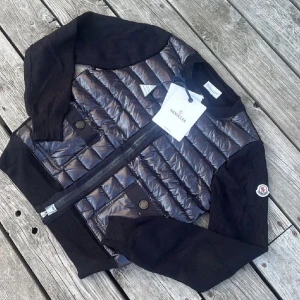 Svart quiltad bomberjacka från Moncler - Snygg svart bomberjacka från Moncler med quiltad glansig front och två fickor med knapp. Ärmarna är stickade och har ribbade muddar. Jackan har dragkedja framtill och klassisk Moncler-logga på ärmen. Perfekt för dig som vill ha en stilren och exklusiv look.