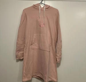 Rosa hoodieklänning från Gant - Mysig rosa hoodieklänning från Gant med broderad logga och känguruficka framtill. Klänningen har huva med snörning och långa ärmar. Klänningen är mer rosa i verkligheten, se bild 2.