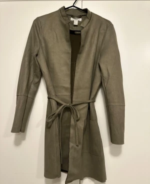 Mörkgrön mockaliknande trenchcoat H&M - Stilren mörkgrön trenchcoat från H&M i mockaliknande material. Jackan har lång ärm, knytband i midjan och två stora fickor framtill. Enkel krage utan slag och rak passform ger en clean look. 