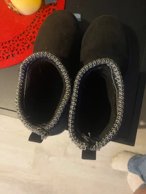 Svarta mockaboots med stickad kant - Svarta boots i mocka med platt sula och rund tå. Skorna har en snygg stickad kant i grått och svart runt öppningen och en praktisk ögla bak för enkel påtagning. Perfekta för kalla dagar och riktigt sköna att ha på sig.