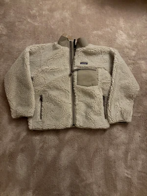 Beige teddyjacka från Patagonia - Säljer en beige teddyjacka från Patagonia med mjukt och fluffigt fleece-material. Jackan har hög krage, dragkedja framtill, två sidofickor med dragkedja och en bröstficka i kontrasterande tyg. Perfekt för kalla dagar och chill stil.