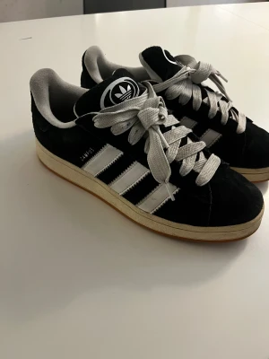 Adidas Campus svarta sneakers mocka  - Svarta Adidas Campus sneakers med klassiska vita ränder och logga på tungan och hälen. Skorna har en låg profil, rund tå och platt sula. Ovandelen är i mjuk mocka med vita snören och detaljer i vitt skinn. Perfekt för dig som gillar retrostil och streetwear.