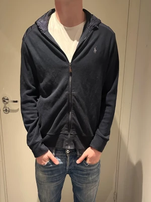 Mörkblå cardigan från Ralph Lauren - Snygg mörkblå cardigan från Ralph Lauren med dragkedja och huva. Tröjan har långa ärmar, ribbade muddar och klassisk Polo-logga på bröstet. Materialet är mjuk bomull och huvan har en snygg kontrast i tyg. Perfekt för chill dagar och streetwear.