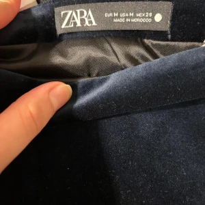Mörkblå sammet kjol från Zara - Snygg mörkblå kortkjol från Zara i mjuk sammet. Kjolen har en rak och enkel design utan mönster eller detaljer, vilket gör den lätt att matcha med olika stilar. Perfekt för dig som gillar klassiska och stilrena plagg.