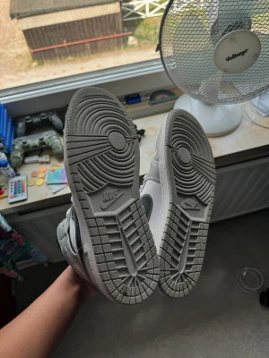 Nike Air Jordan 1 Mid grå/vit - Säljer ett par Nike Air Jordan 1 Mid sneakers i grått och vitt med svarta detaljer. Skorna har klassisk snörning, perforerad tå och rund tå. Swoosh-loggan i svart på sidan och Air Jordan-märke på ankeln. Tillverkade i skinn och syntetmaterial för en clean och sportig look.