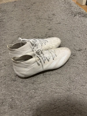 Vita adidas fotbollsskor med snörning - Säljer ett par vita adidas fotbollsskor med snörning och låg profil. Skorna har en rund tå och platt sula med dobbar för gräsplan. Ovandelen är i syntetmaterial med diskreta mönster och adidas-logga på hälen. Perfekt för dig som vill ha stilrena och lätta skor på planen. Tveka inte att höra av er vid några frågor eller funderingar🤗