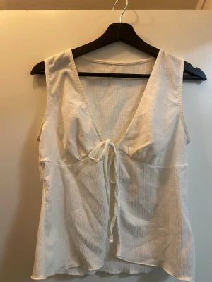Vit omlottopp med knyt strl S - Från lager 157. Säljer en vit ärmlös omlottopp med djup v-ringning och knyt framtill. Toppen har en lätt strukturerad yta och är tillverkad i ett mjukt, tunt tyg som passar perfekt till varma dagar. Enkel och stilren design som är lätt att matcha.