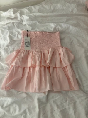 Rosa volangkjol från Neo Noir - Supersöt kortkjol från Neo Noir i färgen Rose Smoke. Kjolen har smockad midja och dubbla volanger som ger en flowy och feminin look. Perfekt att styla med en enkel topp för en trendig vibe. Materialet känns lätt och luftigt.