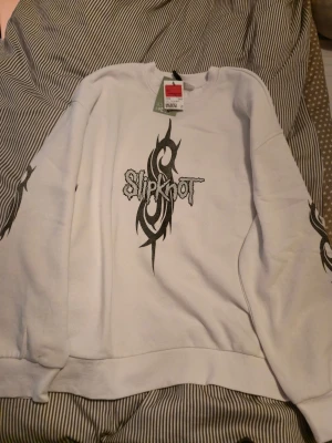 Vit Slipknot sweatshirt - Vit sweatshirt med Slipknot-tryck framtill och tribal-mönster på ärmarna. Tröjan har en klassisk passform med rund hals och långa ärmar. Köpt men ej använd. Tjock och varma, perfekt myströja. Oversized fit