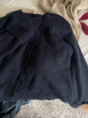 Mörkblå stickad kofta med knappar - Mysig mörkblå stickad kofta med rund hals och knäppning framtill. Koftan har långa ärmar och en enkel, klassisk design som passar perfekt till lager-på-lager-stil under kyliga dagar.